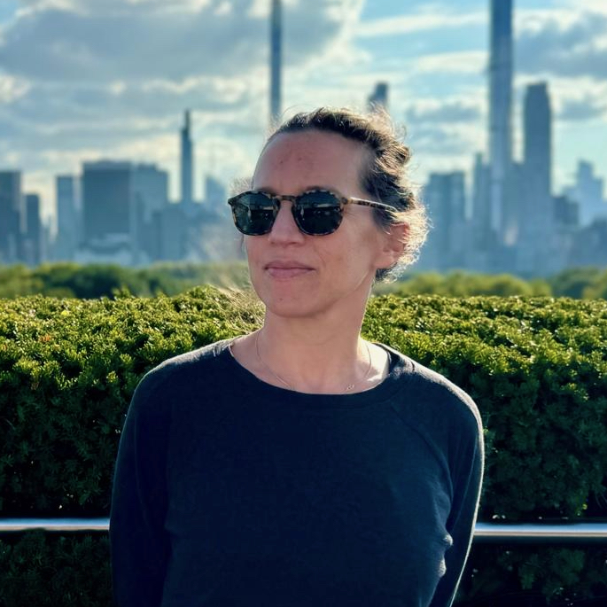 Gabriella Kessler standing outside in front of a bush and a city skyline in a navy blue sweater and black sunglasses